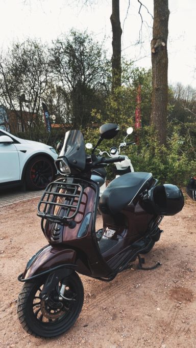 Zwarte scooter met mandje, geparkeerd tussen bomen en een witte auto.